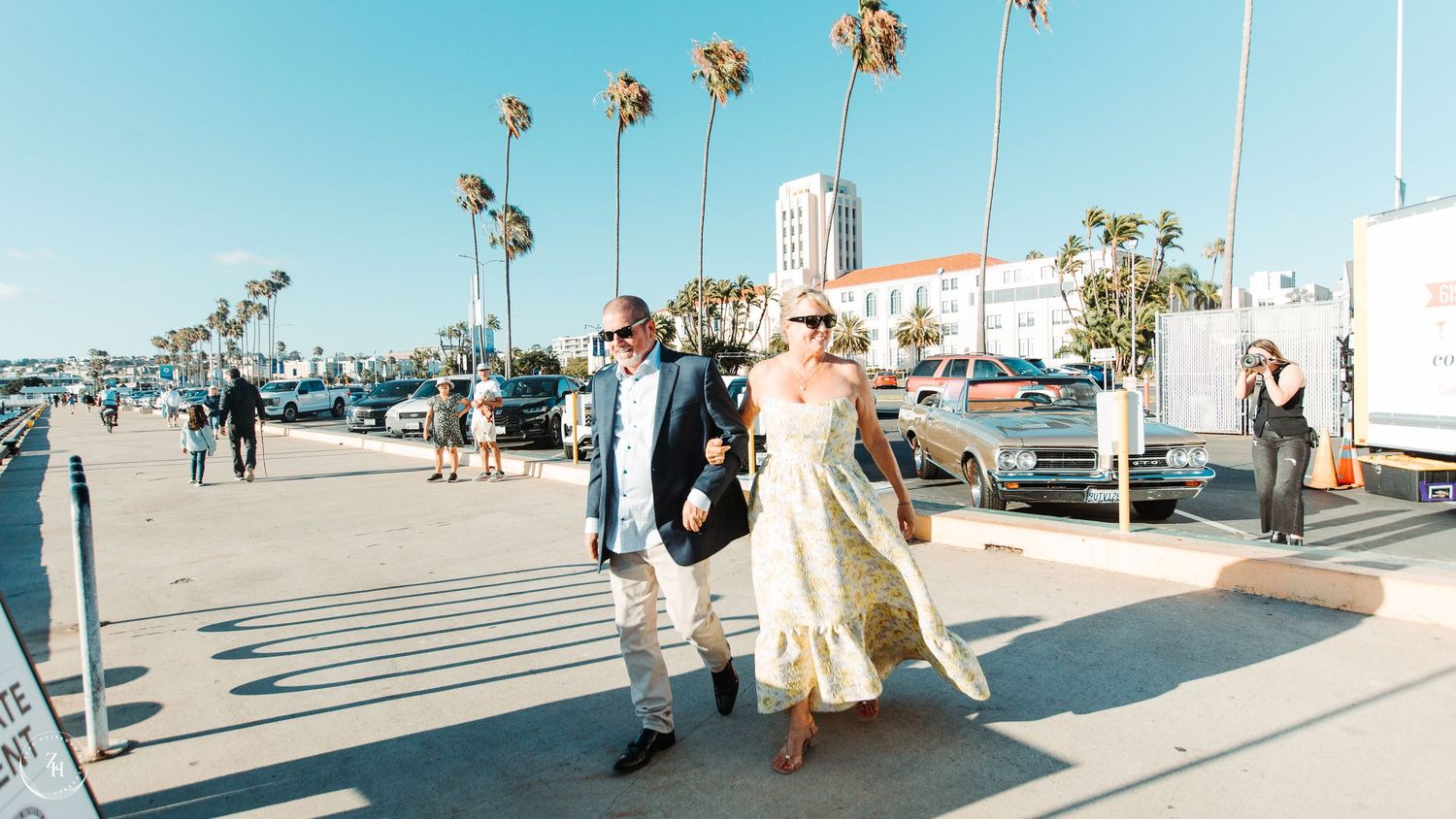 Couple walking hand in hand along sunny Santa Monica boardwalk with palm trees and blue sky.