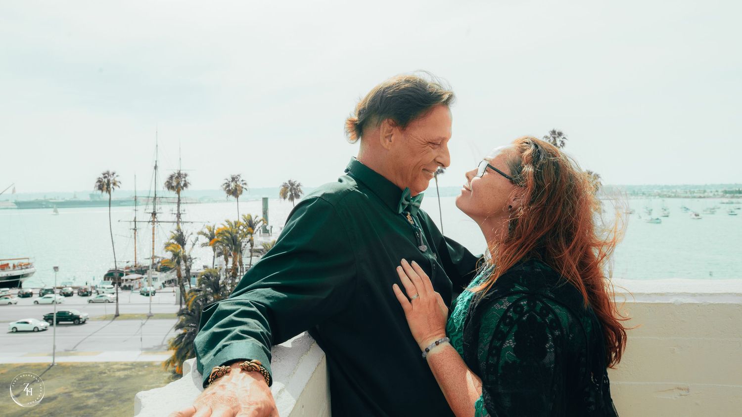 Romantic couple embraces on coastal balcony with ocean and palm trees in background.