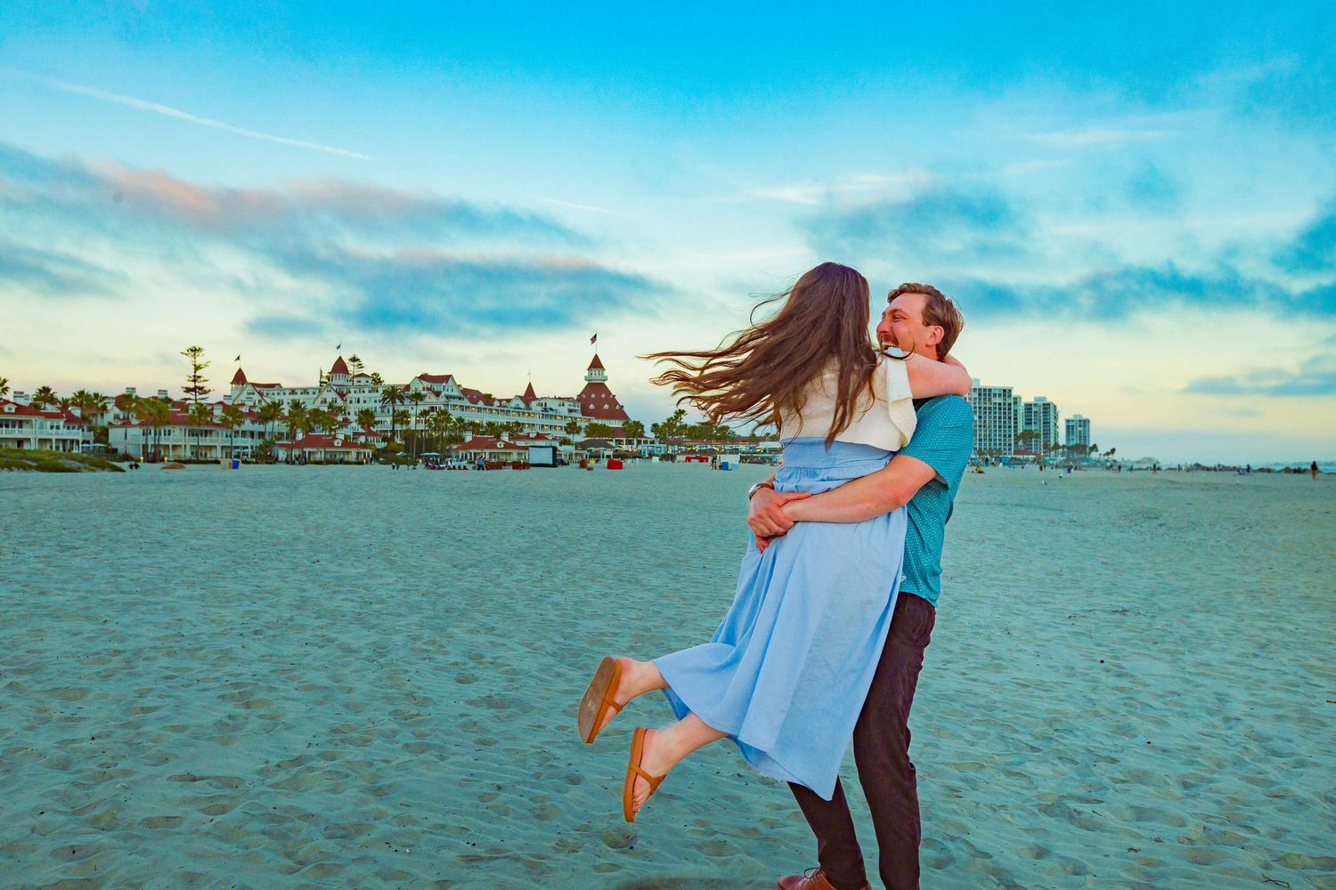A couple embraces romantically on a sandy beach at sunset with a historic hotel and city skyline in the background.