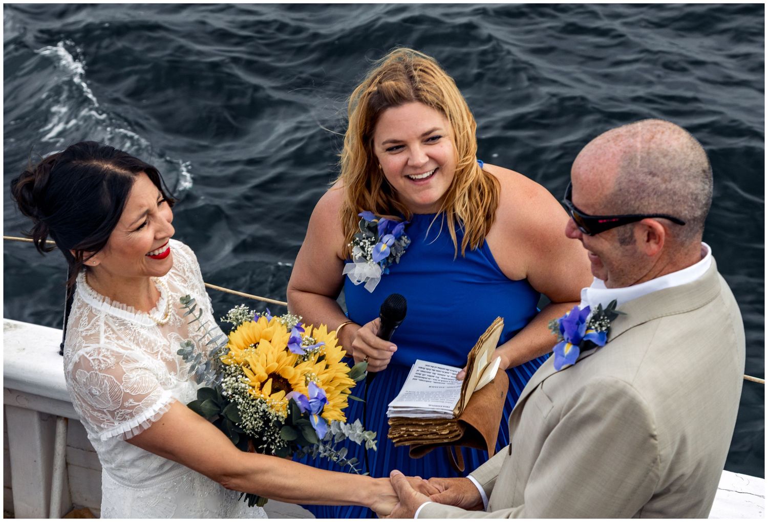 Bride and groom exchange vows on a boat with smiling officiant in blue dress holding sunflower bouquet.