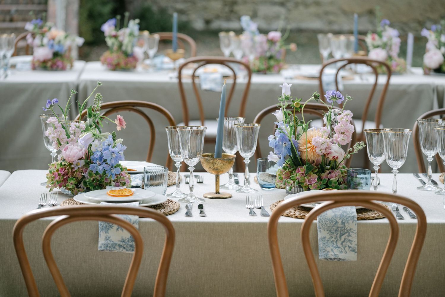 Elegant wedding reception table with bentwood chairs, floral centerpieces in pastel hues, crystal glassware, and a neutral linen tablecloth.