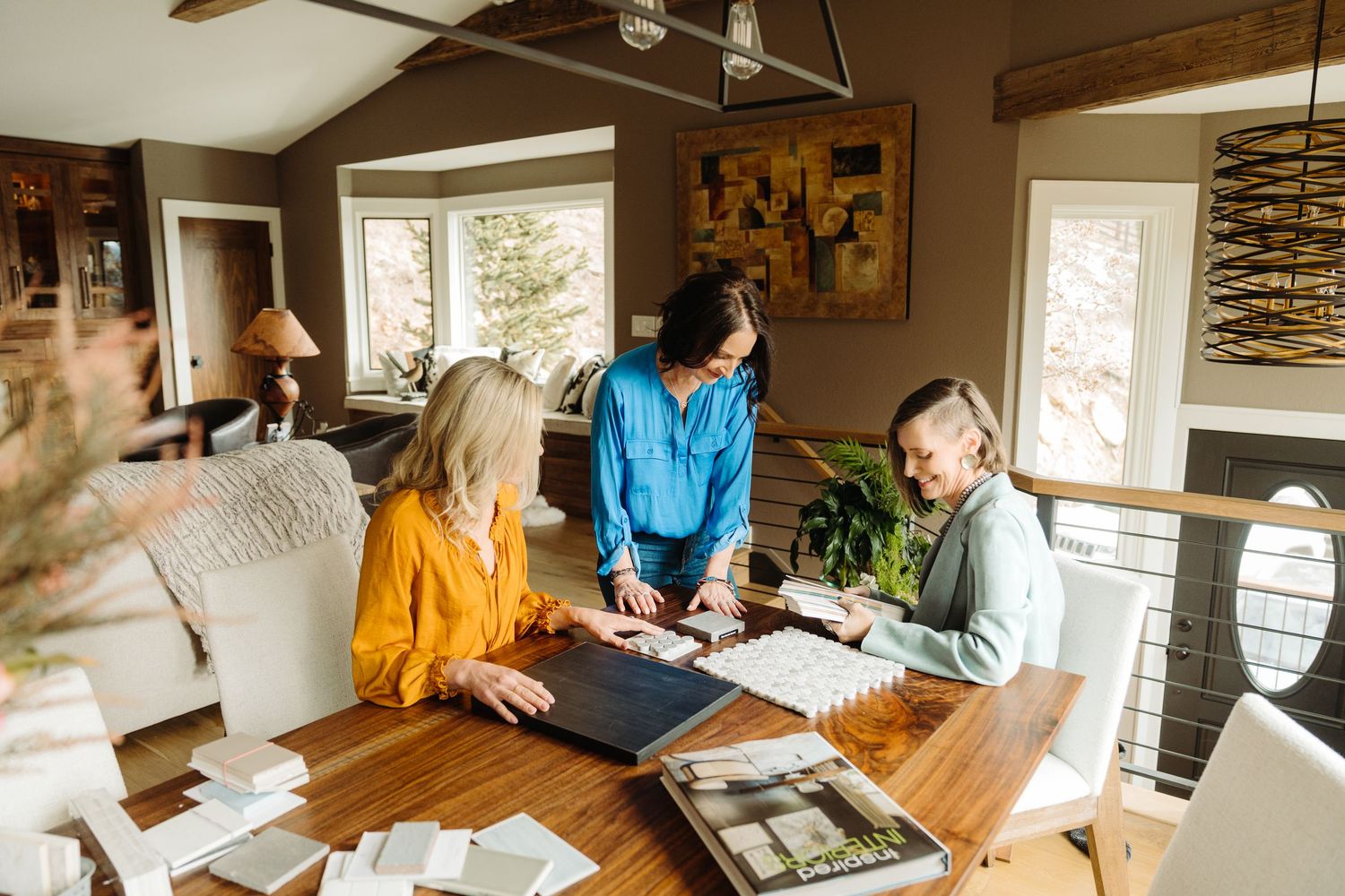 Three women collaborating over design samples and portfolios at a wooden dining table in a modern home.
