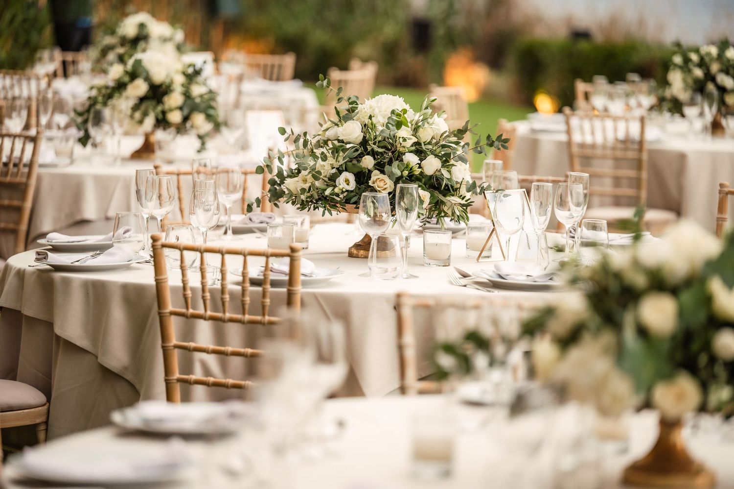 Elegant outdoor wedding reception with ivory linen tables, chiavari chairs, and white floral centerpieces against a lush green backdrop.