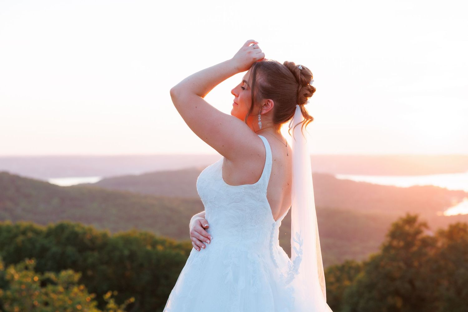 Bride in white gown and veil poses at sunset overlooking scenic mountain and lake landscape.