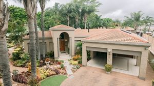 Aerial view of a luxury home with terracotta roof, manicured gardens, palm trees, and a double garage in a tropical setting.