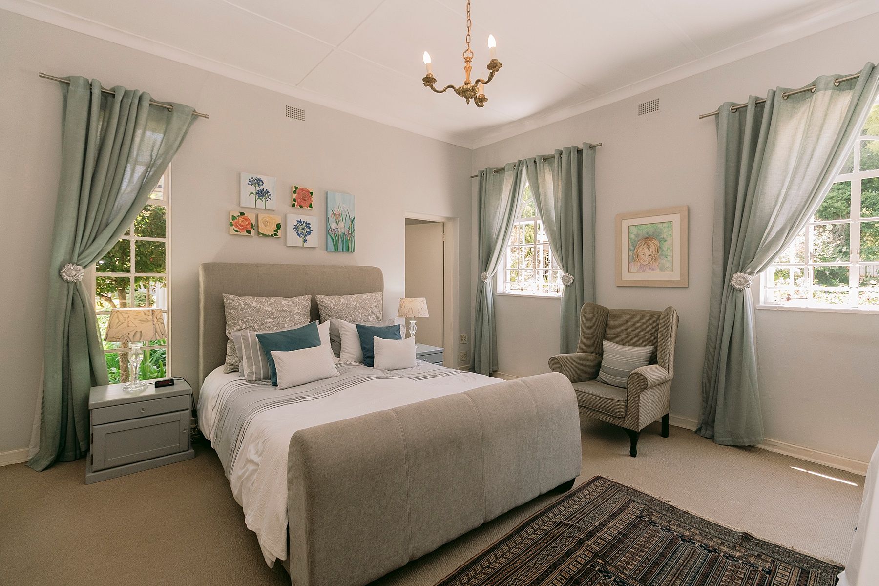 Elegant bedroom with sage green curtains, gray upholstered bed, chandelier, and natural light from two windows.