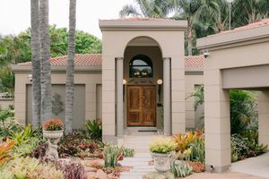 Elegant Mediterranean-style home entrance with arched doorway, wooden double doors, lush tropical landscaping, and palm trees.