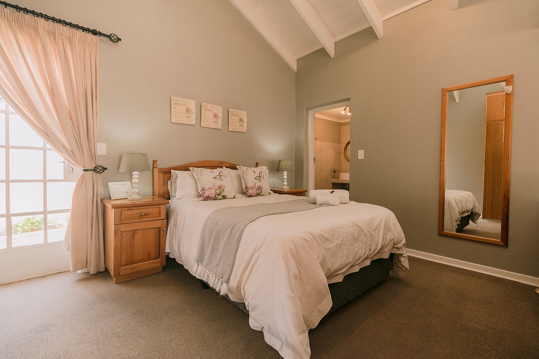 Cozy bedroom with a wooden bed, neutral decor, vaulted ceiling, large mirror, and natural light from a window.