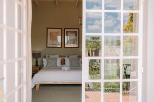 Bright bedroom viewed through open French doors, featuring a neatly made bed with decorative pillows and lush garden views.