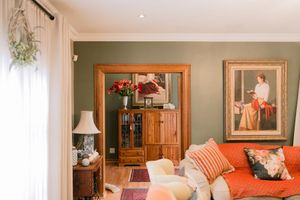 Cozy living room with sage green walls, wooden cabinet, large portrait painting, and orange sofa with decorative pillows.