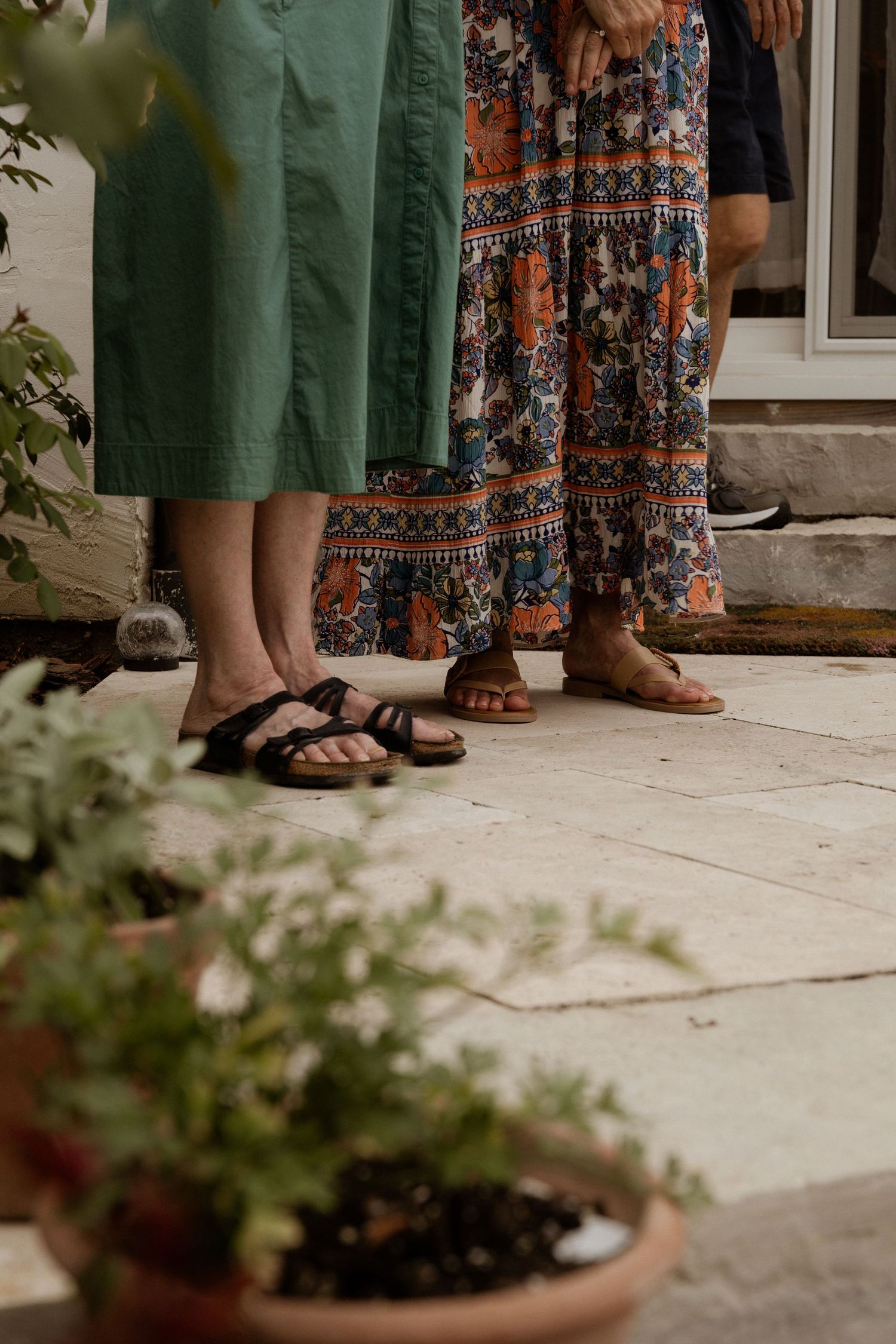 Two women in floral and green skirts wearing sandals stand on a stone patio surrounded by potted plants.