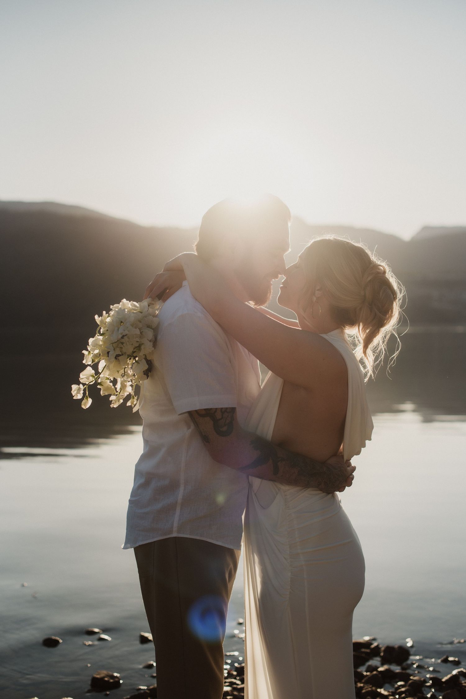Couple sharing a romantic kiss at golden hour by a lake, bride in white gown holding bouquet with sun flare.