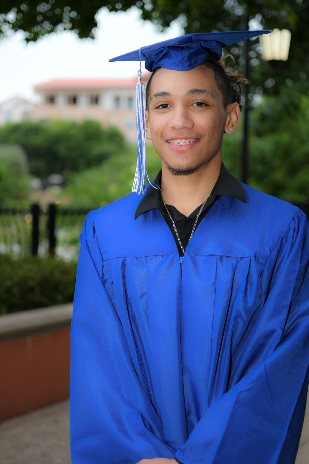 Young man smiling in blue graduation cap and gown outdoors on a sunny day.