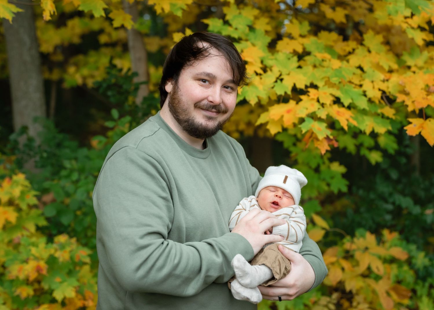 Man in green sweatshirt holding newborn baby outdoors among vibrant yellow and green autumn foliage.