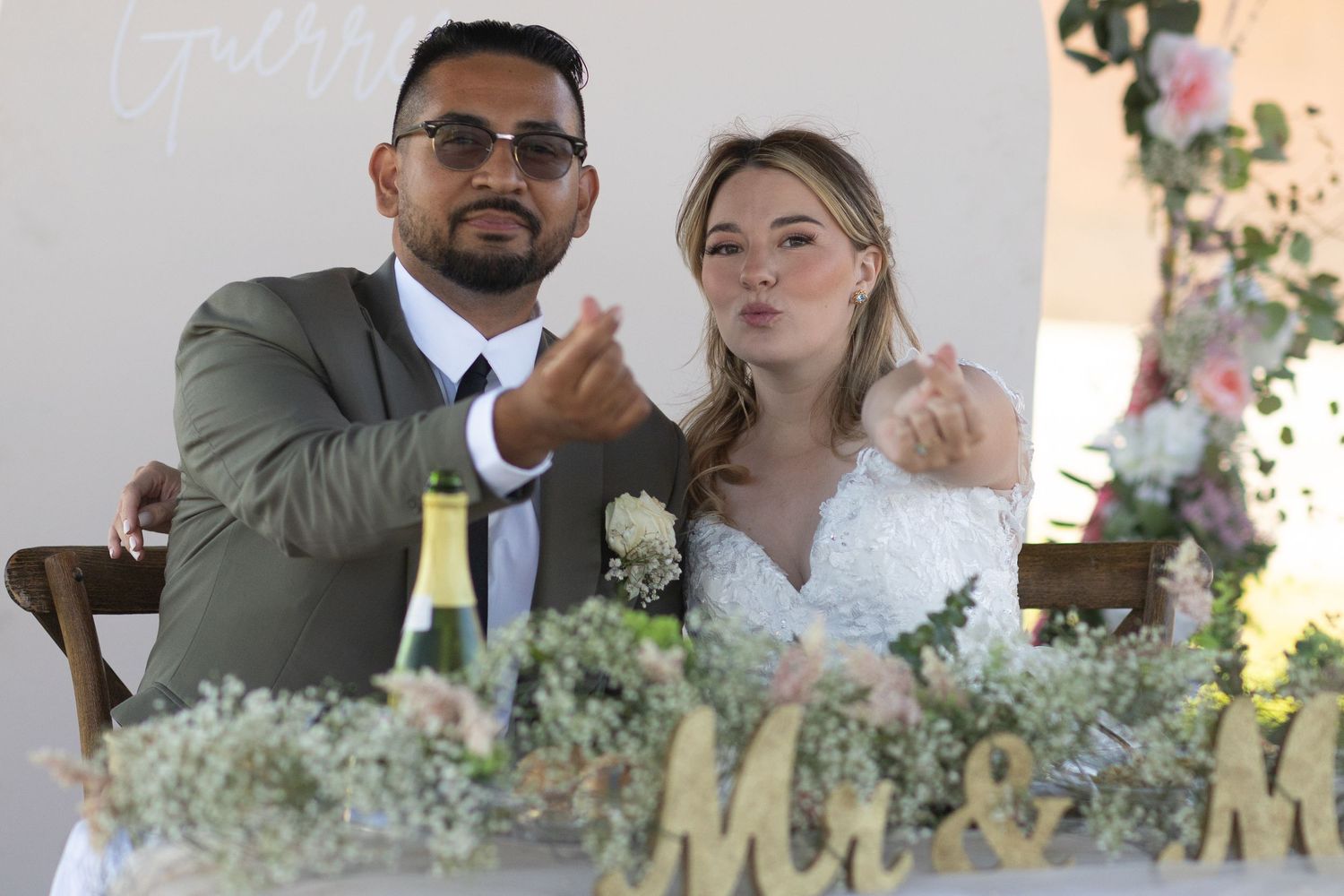 Newlywed couple at wedding reception table with Mr & Mrs sign, champagne, and floral arrangements.