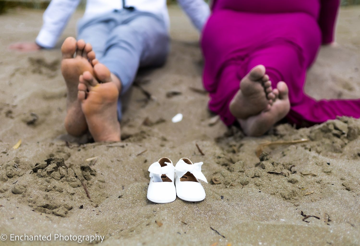 Baby shoes placed on the sand with expectant parents softly blurred in the background