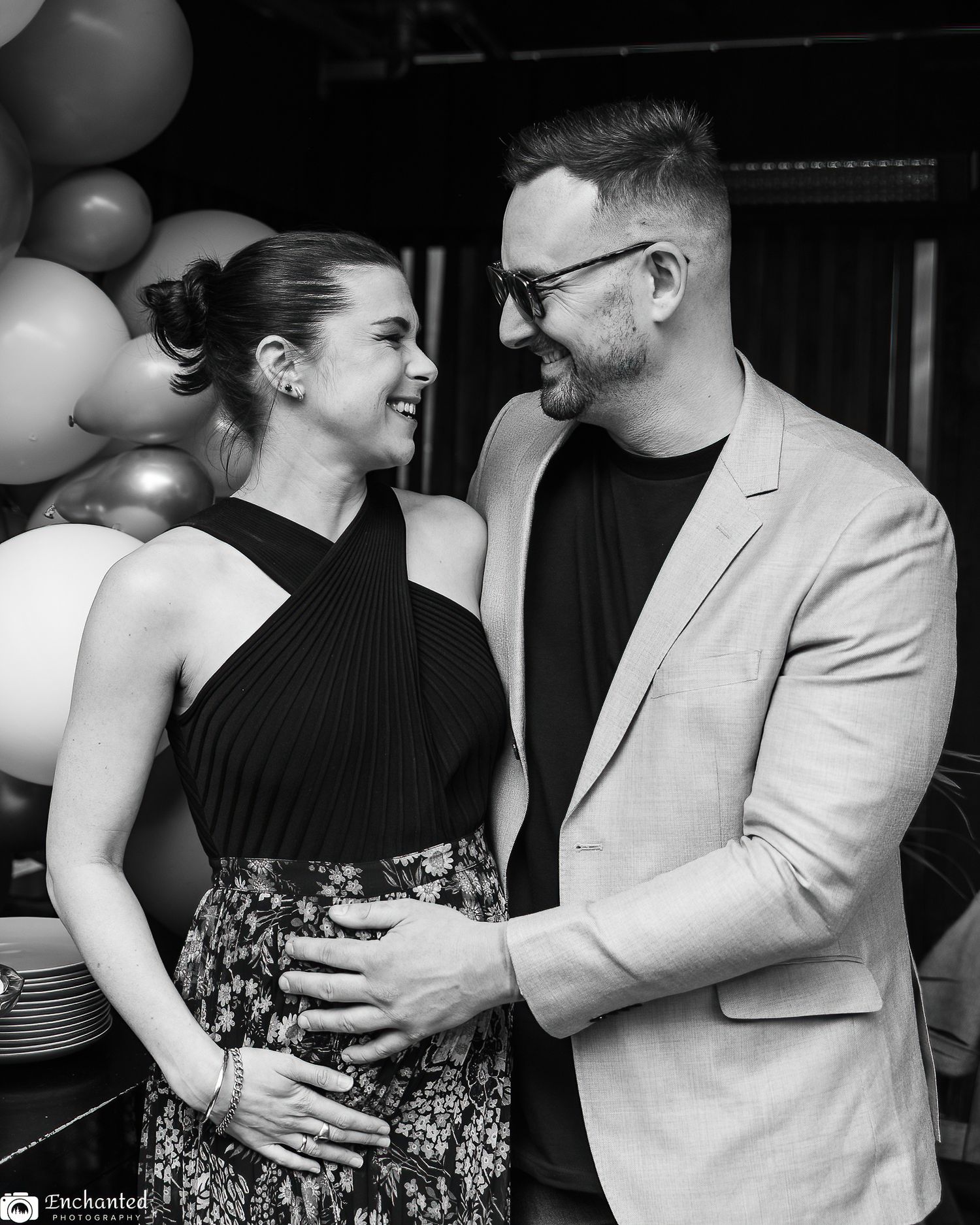 Black and white maternity portrait of a couple smiling at each other while holding the baby bump