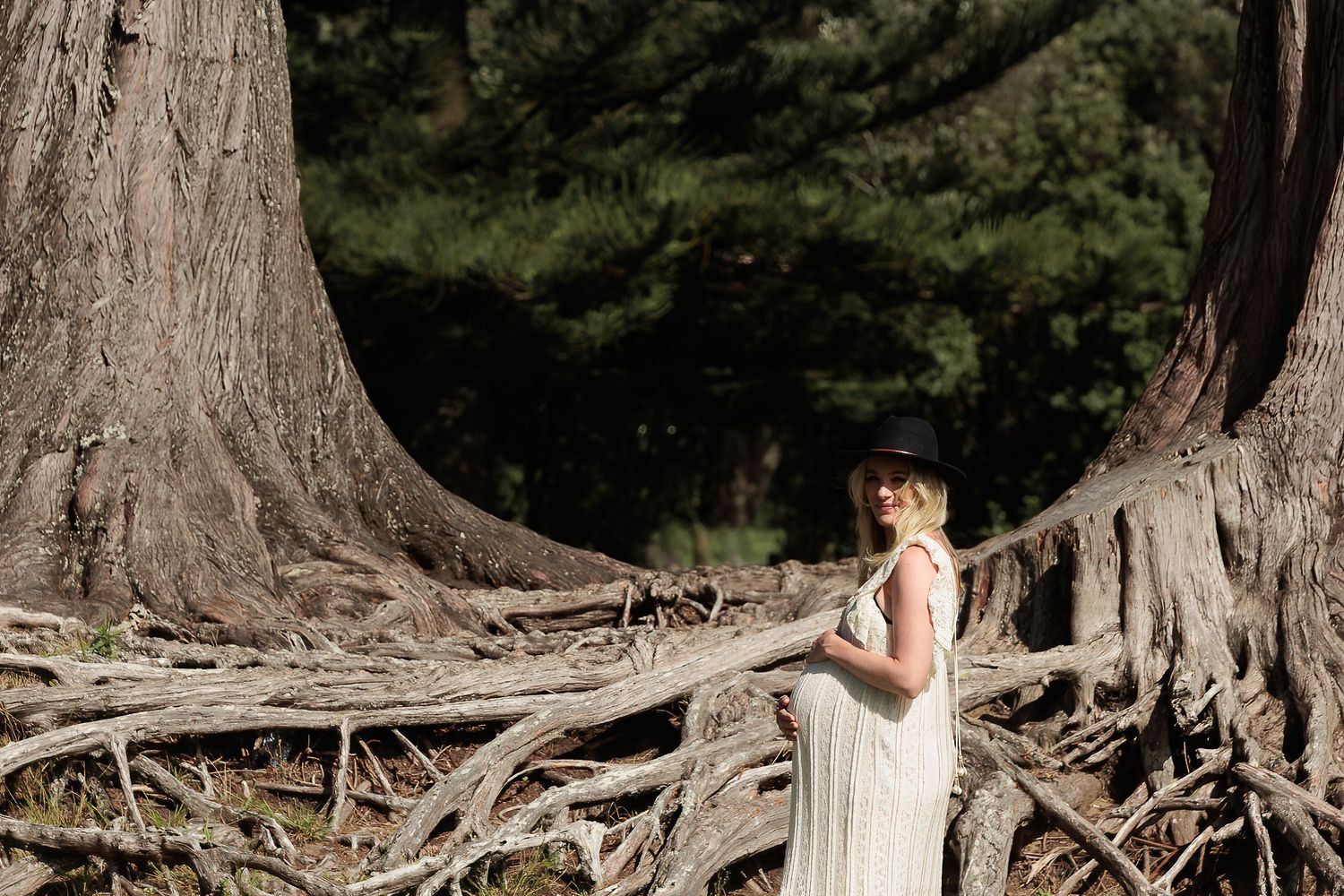 Pregnant woman in a white dress sitting among large tree roots during an outdoor maternity portrait