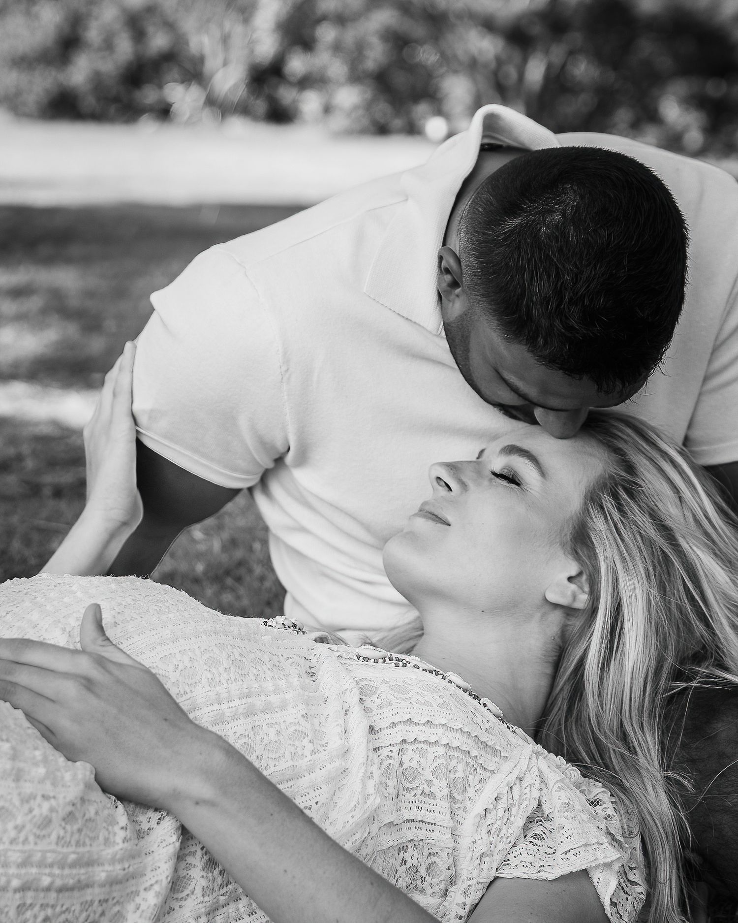 Black and white maternity portrait of a partner gently kissing the expectant mother as she lies on the grass