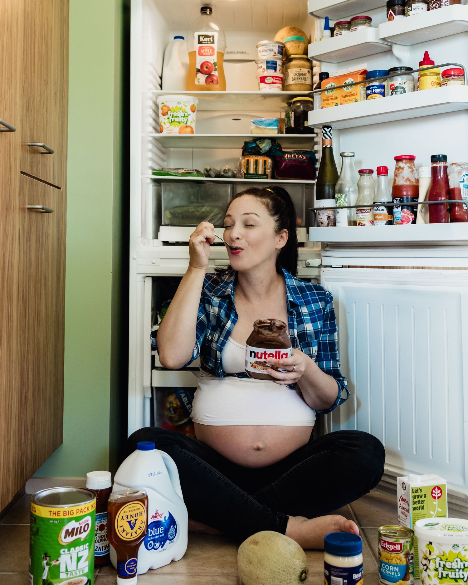 Humorous maternity portrait of an expectant mother sitting on the floor beside an open fridge