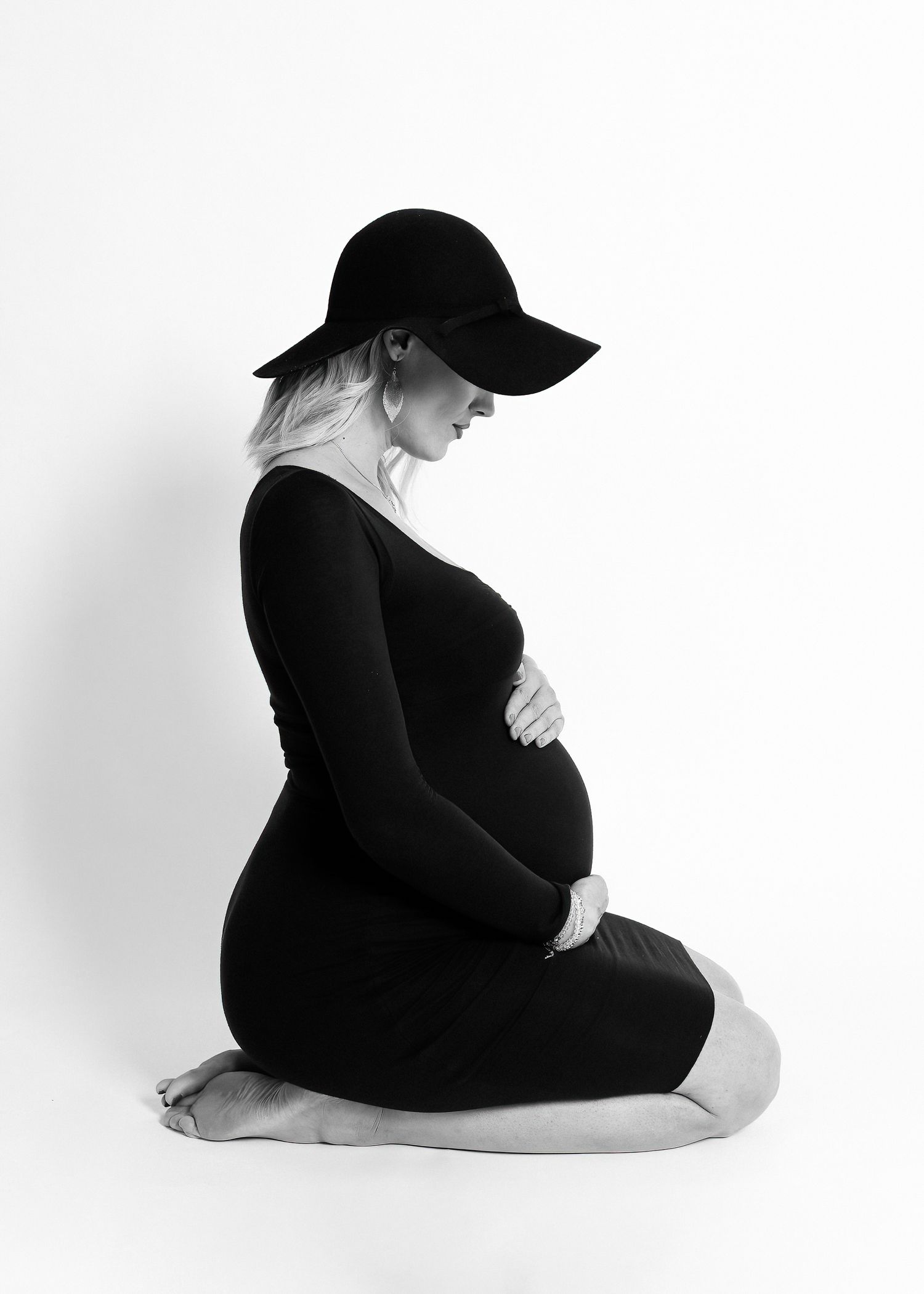 Black and white side-profile studio portrait of a pregnant woman in a fitted dress and wide-brim hat