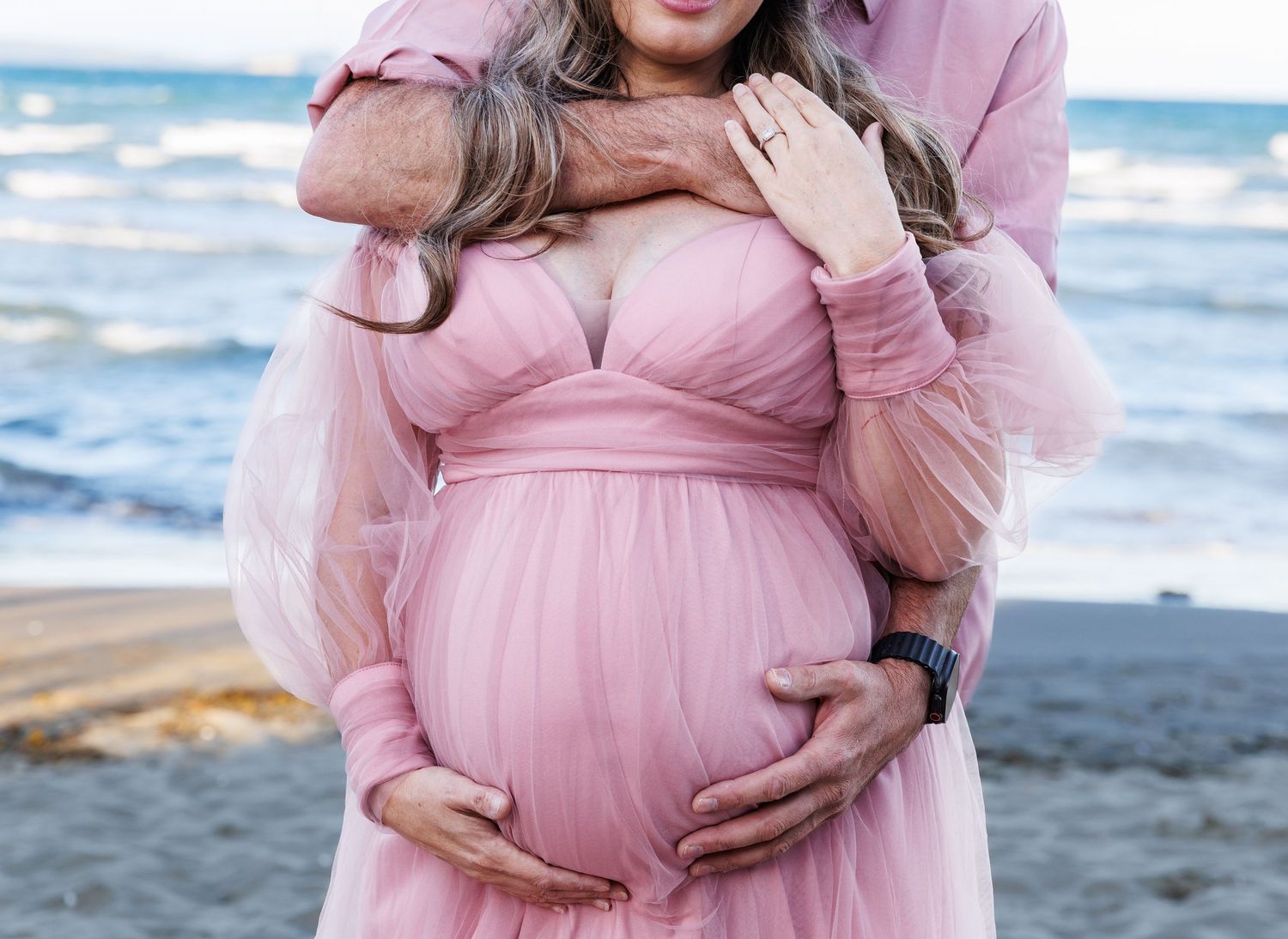 Close-up maternity portrait of a pregnant woman in a pink gown embraced by her partner at the beach