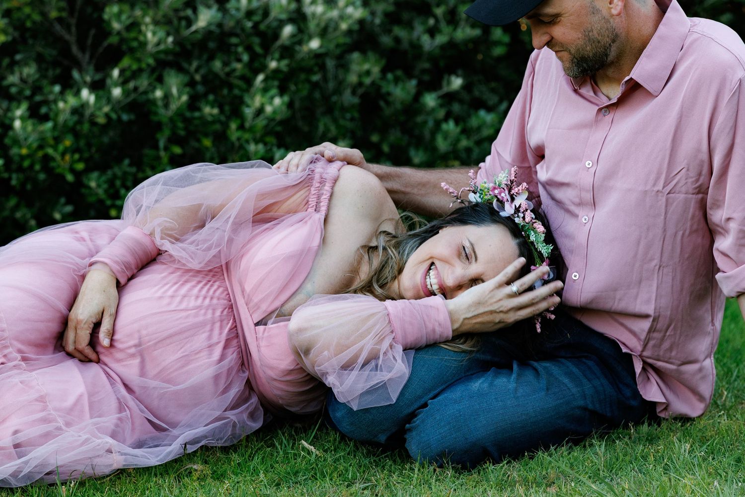 Expectant mother in a pink tulle gown resting across her partner’s lap during an outdoor maternity session