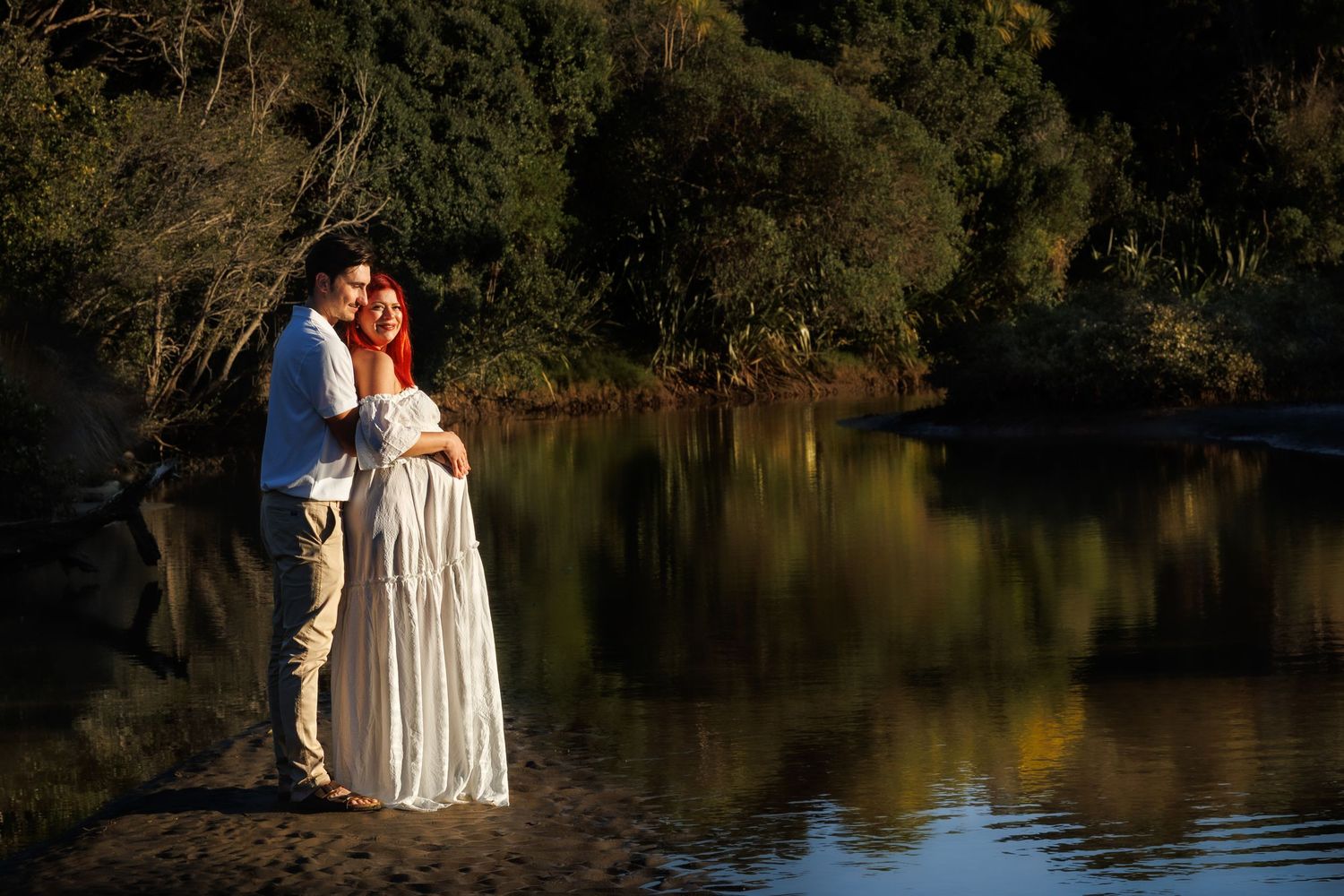 Expectant couple embracing beside calm water during an outdoor maternity session