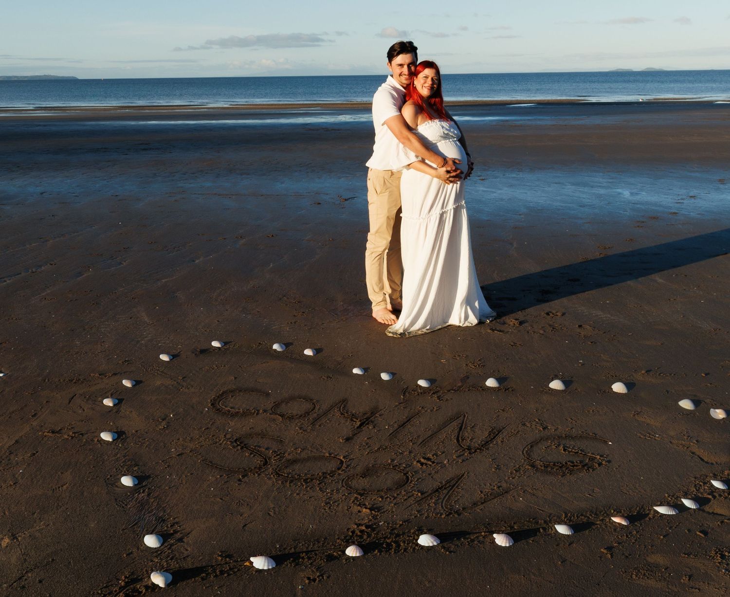 Expectant couple embracing on the beach behind the words “Coming Soon” written in the sand and framed with seashells