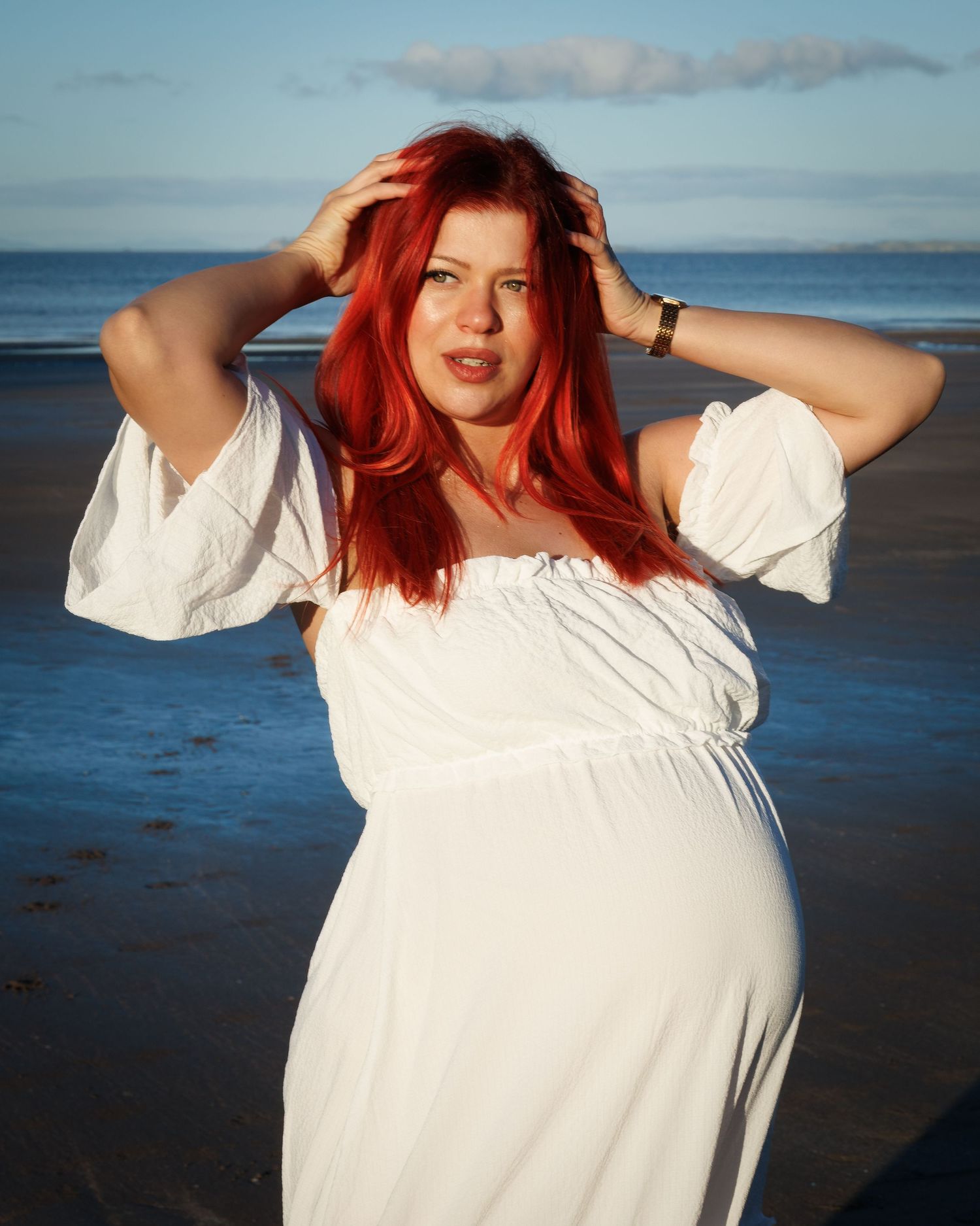 Pregnant woman in a flowing white dress posing on the beach with her hands in her hair