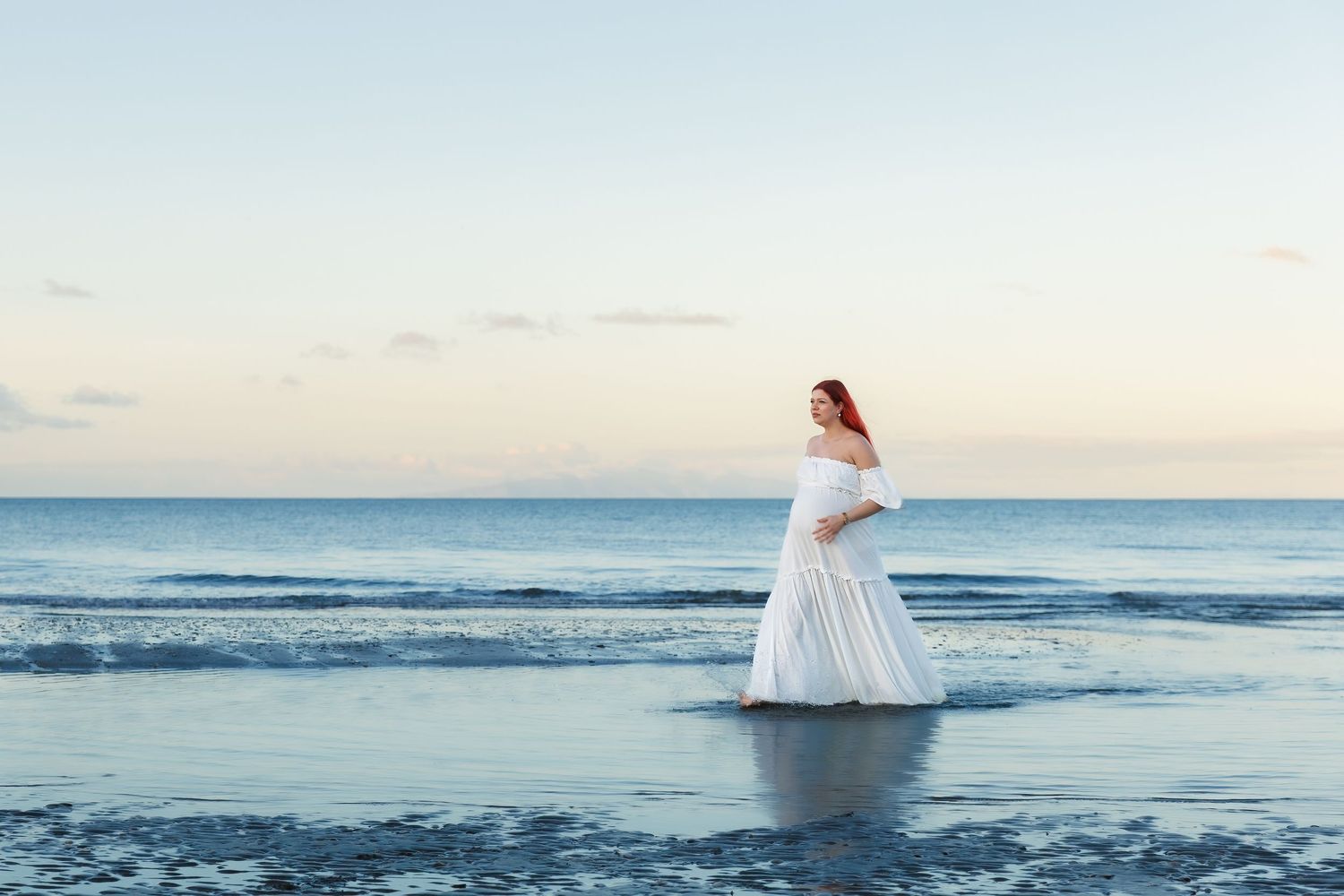 Pregnant woman in a flowing white dress walking in shallow water at an Auckland beach