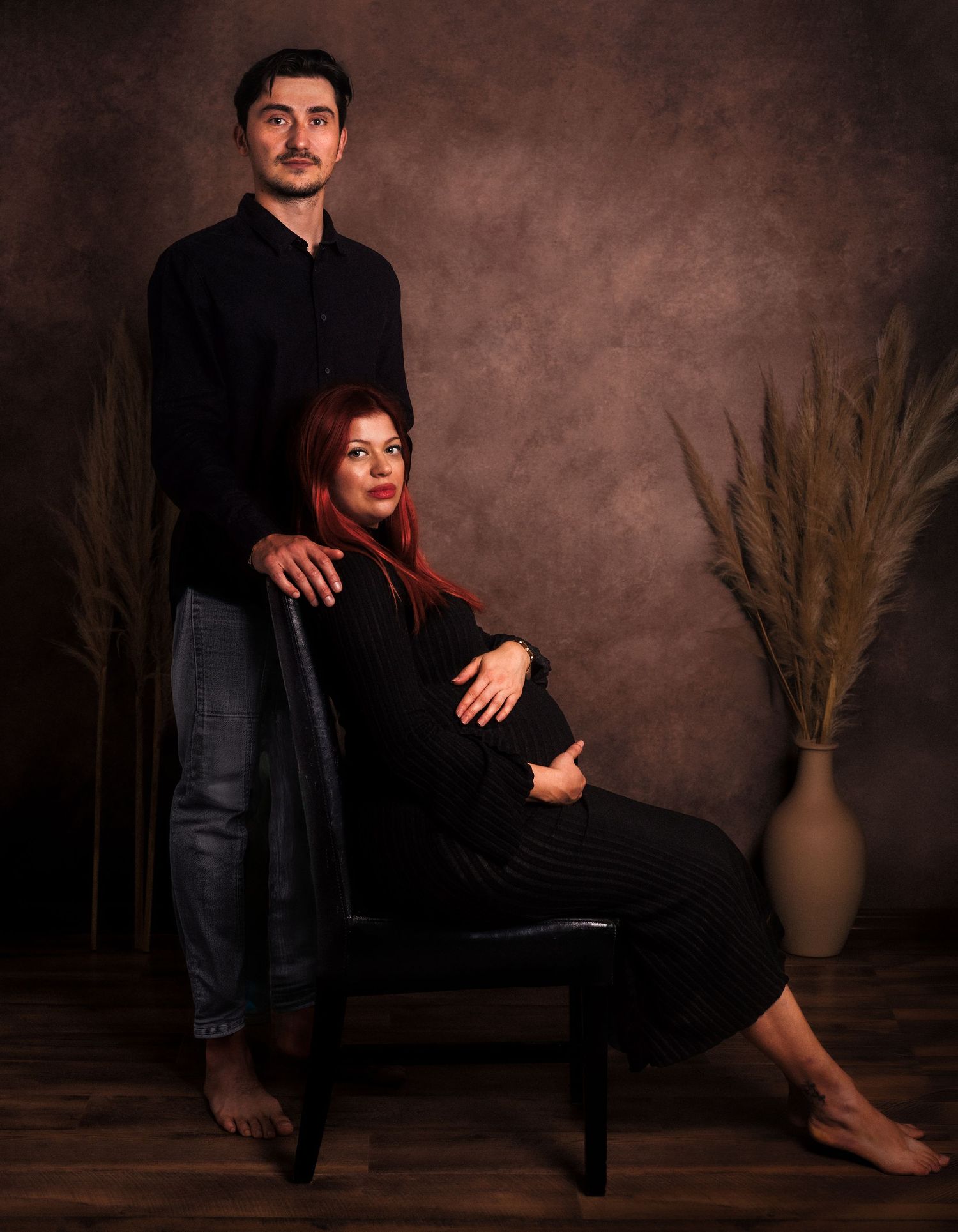 Studio maternity portrait of an expectant mother in a black dress seated beside her standing partner