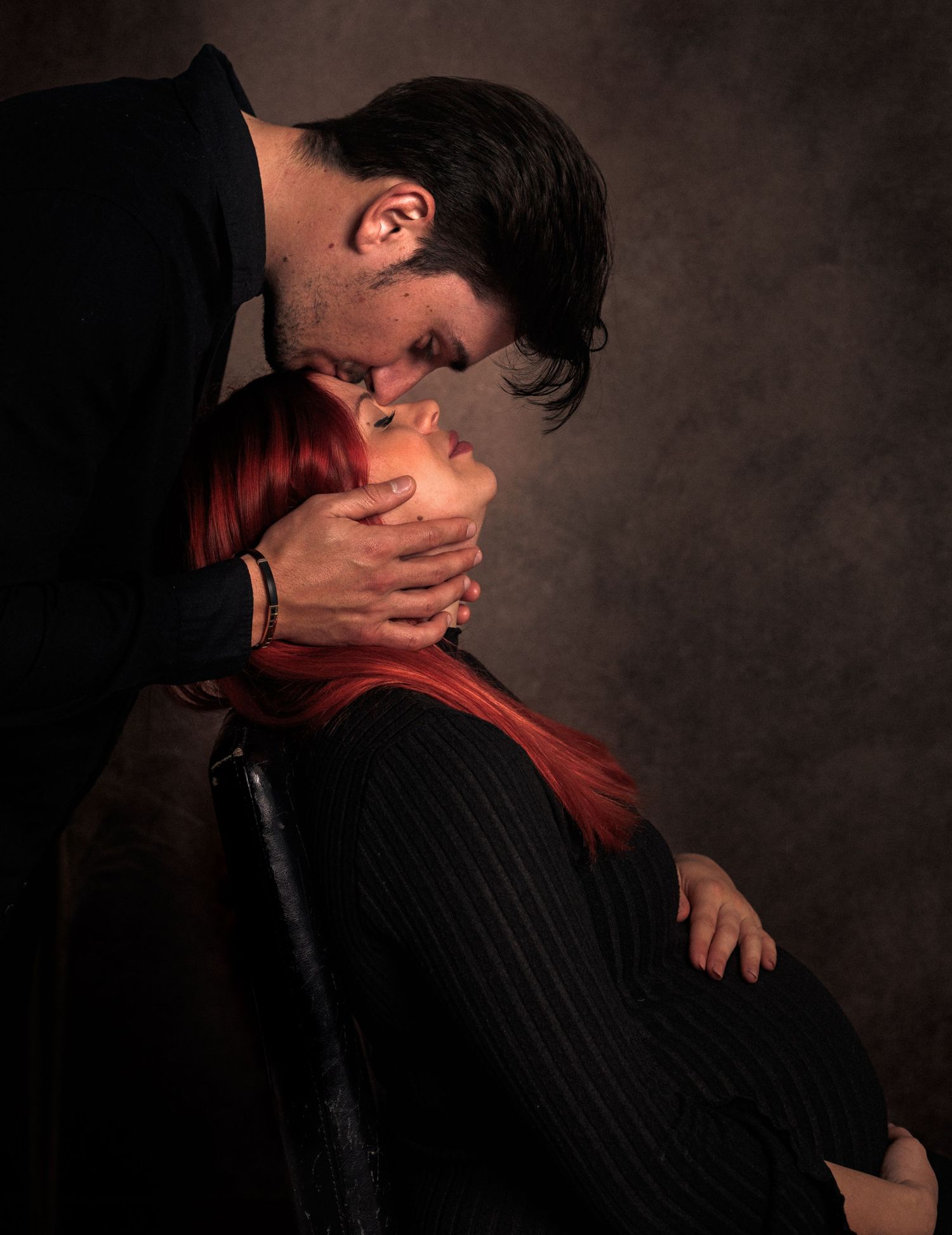 Moody maternity portrait of a seated expectant mother with her partner leaning down to kiss her forehead
