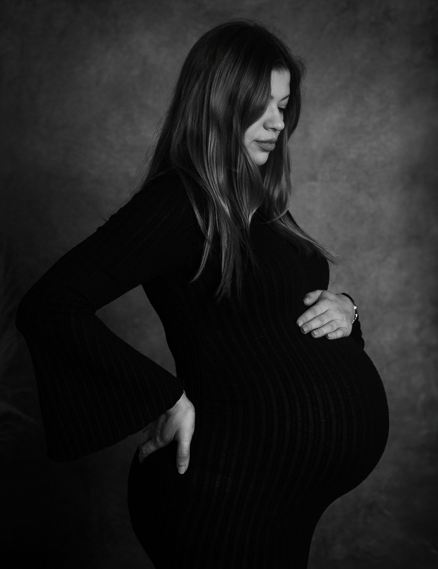 Black and white side-profile studio maternity portrait of a woman in a black dress cradling her bump
