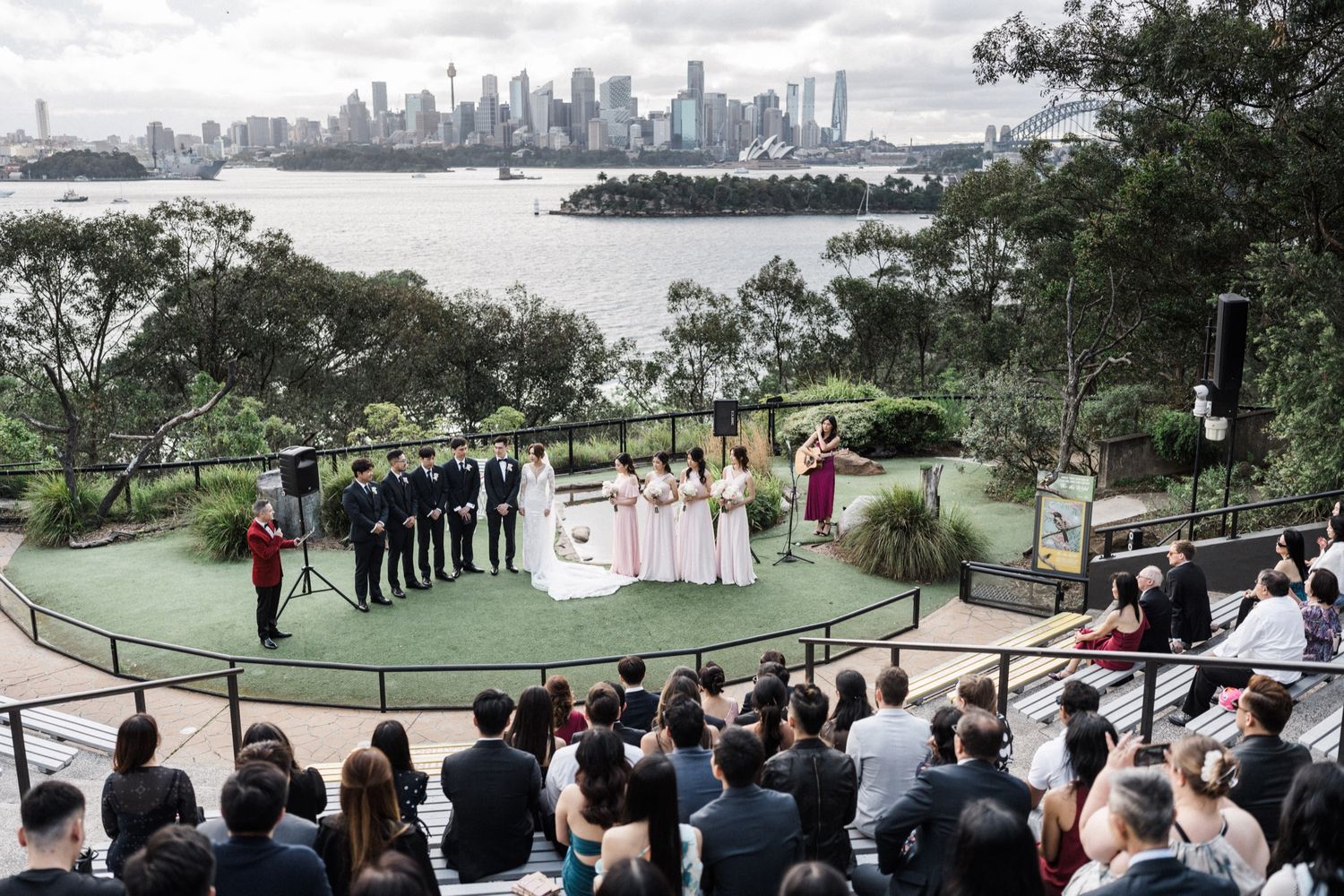 Outdoor wedding ceremony at Taronga Zoo Sydney with city skyline and harbor views, guests seated watching couple exchange vows.