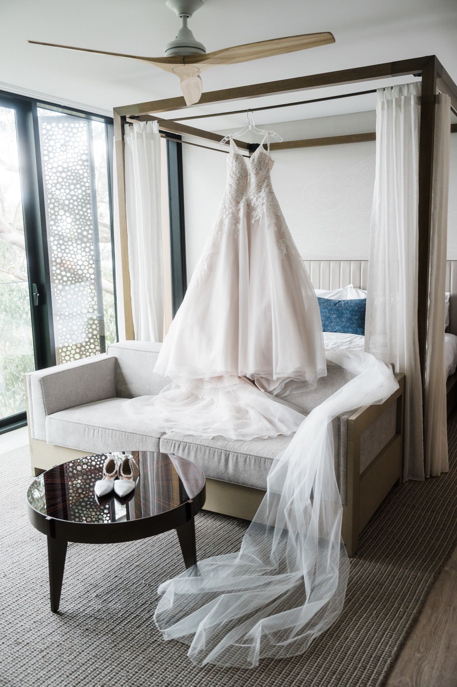 Elegant wedding dress hanging on a four-poster bed with veil draped over, bridal shoes displayed on a dark oval table.