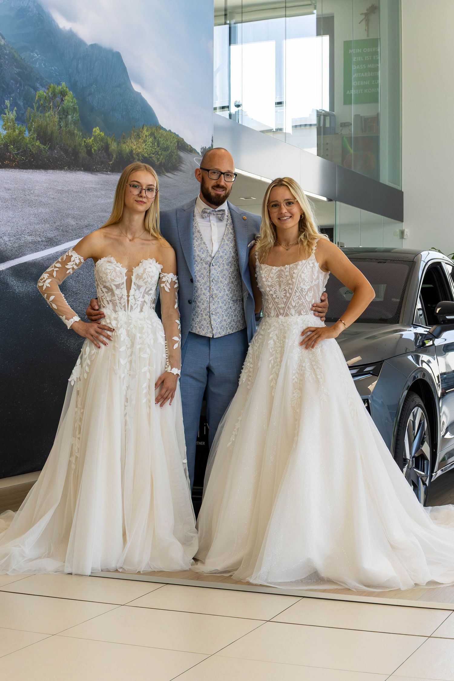 Man in blue suit poses with two women in elegant white lace wedding gowns inside a luxury car showroom.