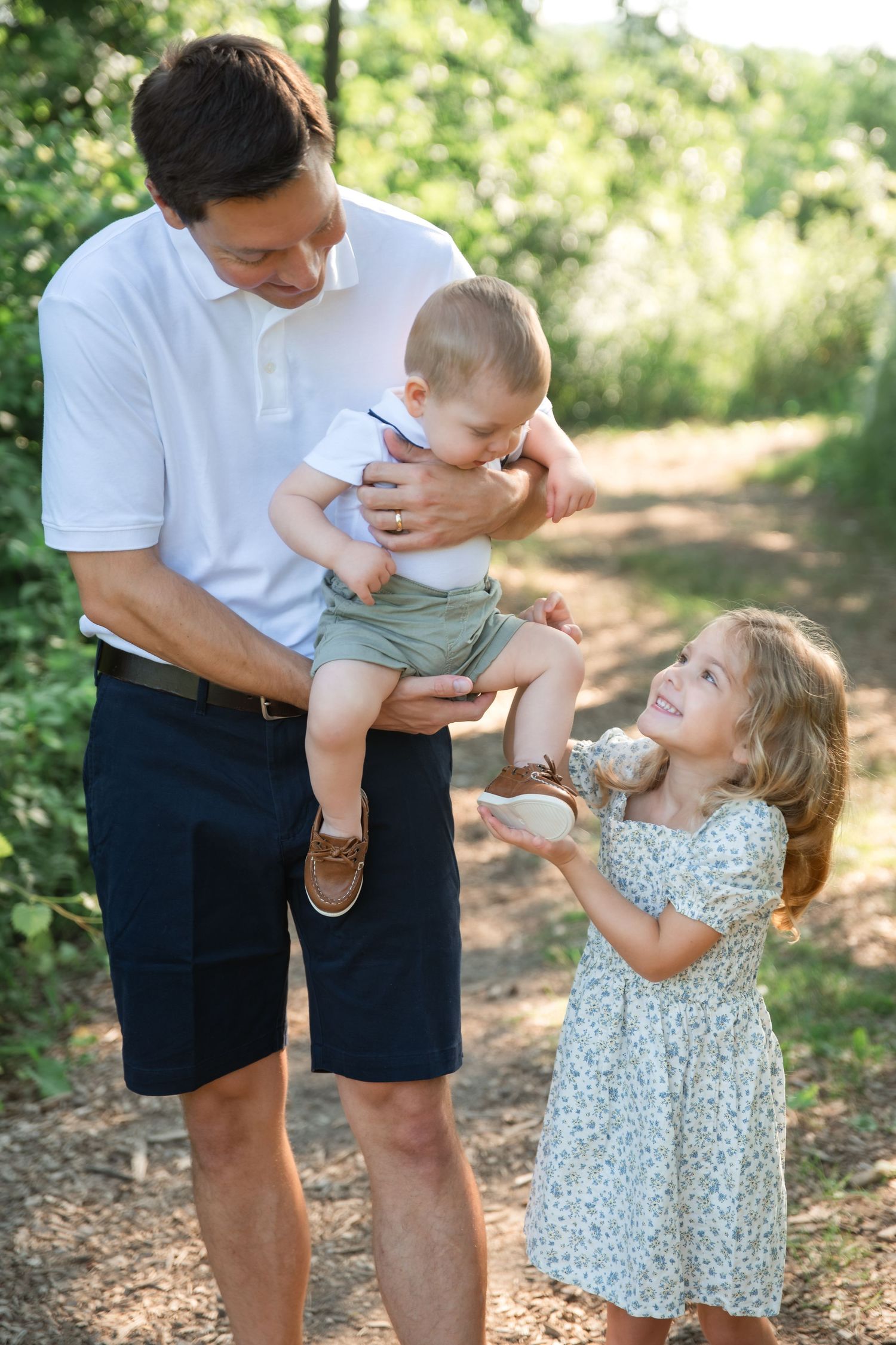 Father holds baby boy while young girl in floral dress smiles up at them on a sunny nature path.
