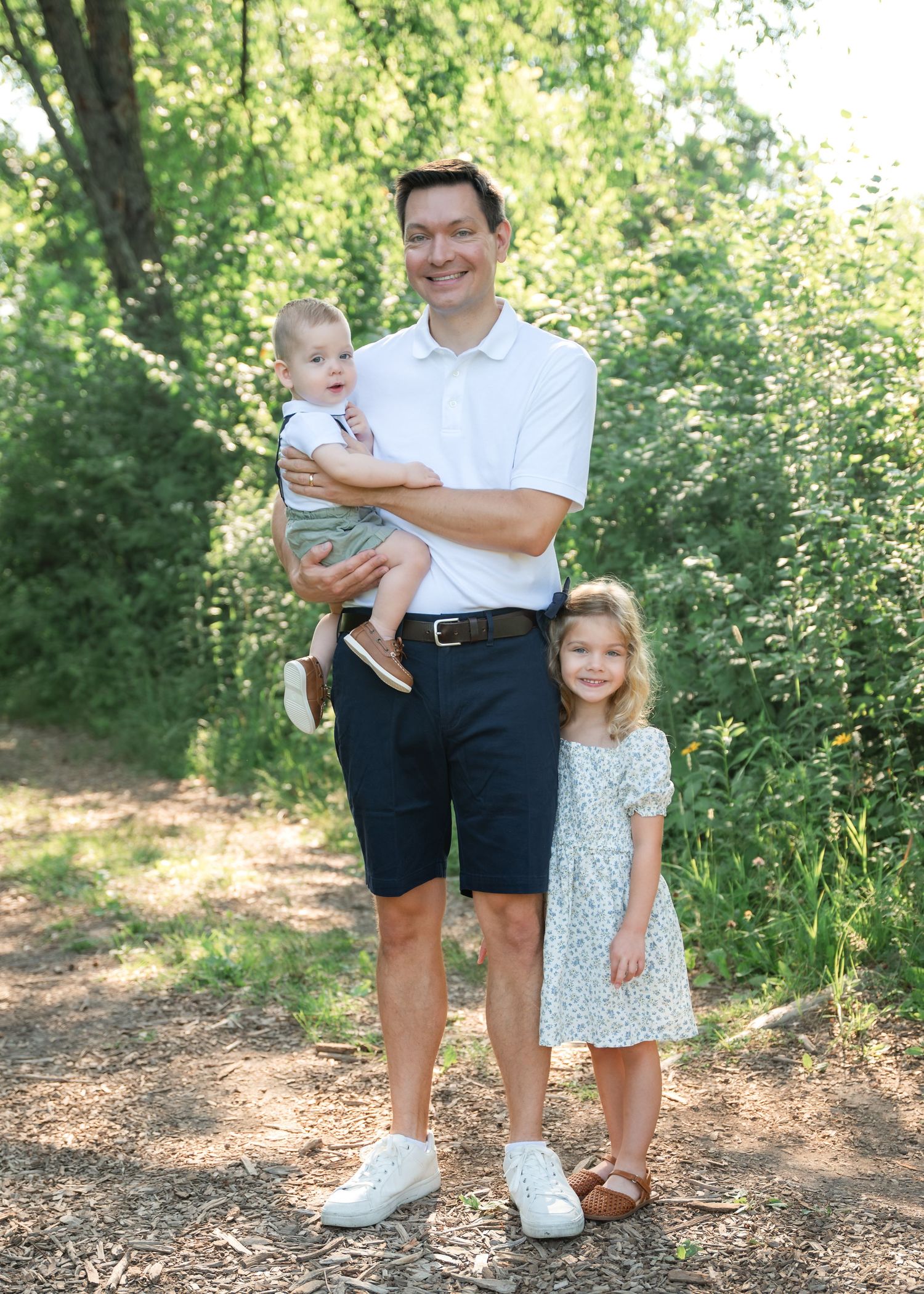 A smiling man holds a baby while standing next to a young girl in a floral dress on a sunny outdoor path.