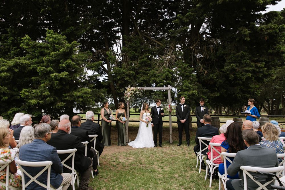 Outdoor wedding ceremony under large trees with bride and groom at altar, guests seated in white chairs on green lawn.