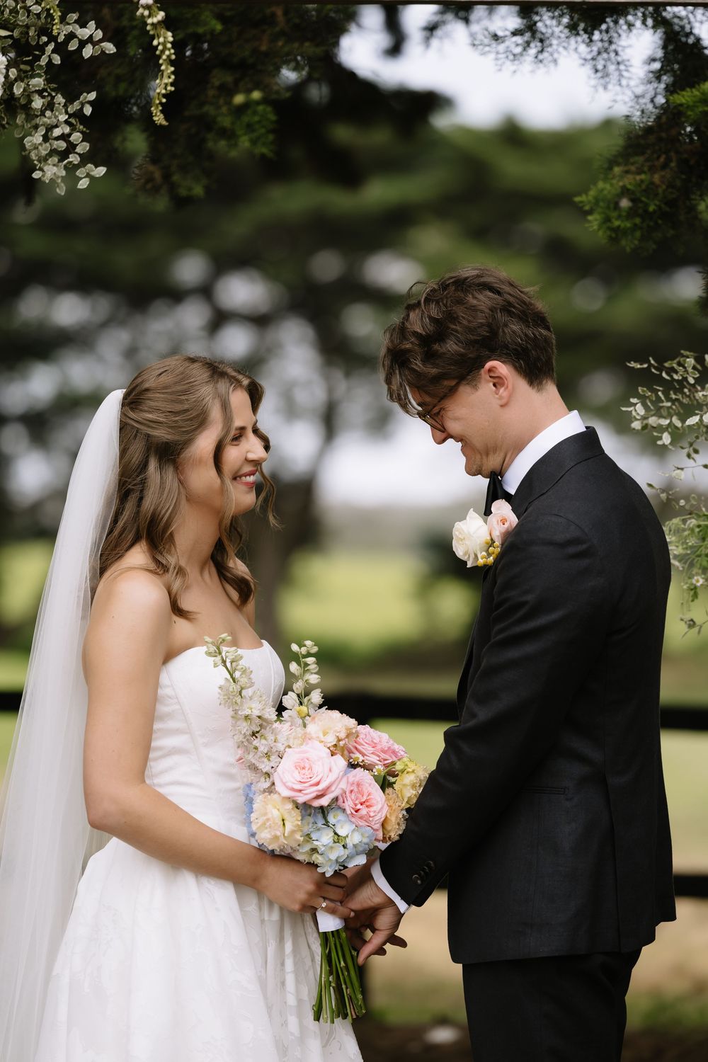Bride and groom holding a pink floral bouquet, smiling at each other outdoors under green foliage on their wedding day.