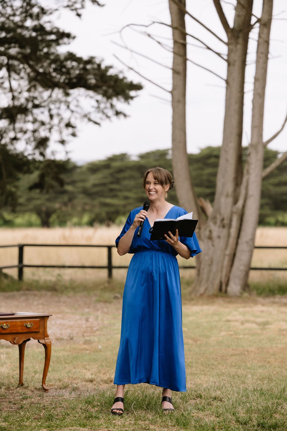 Woman in blue maxi dress holding microphone and book, speaking outdoors at a garden ceremony near trees.