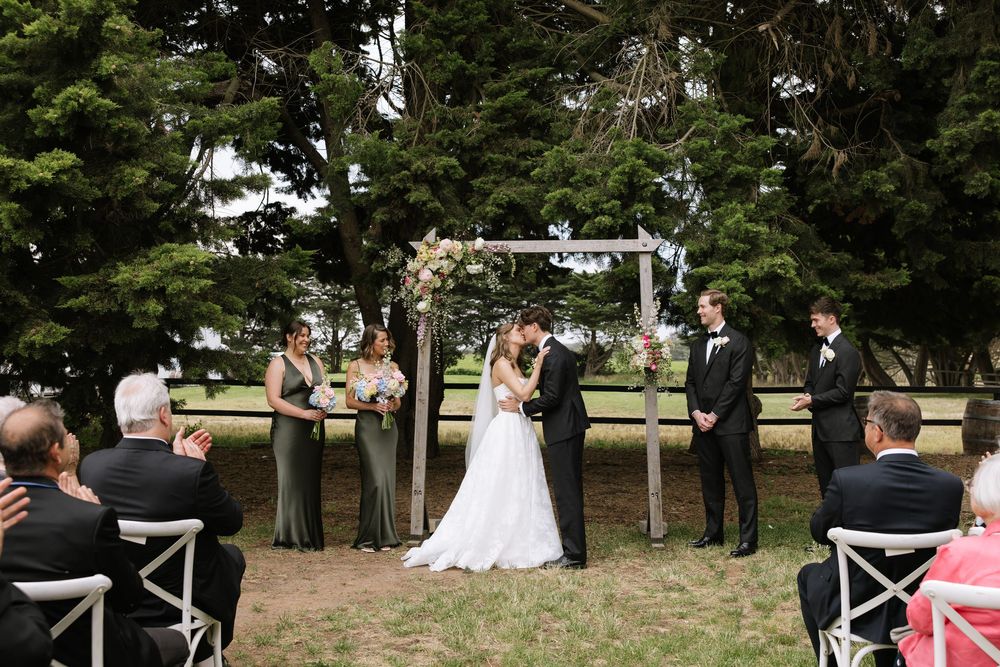 Bride and groom share first kiss at outdoor wedding ceremony under floral arch, surrounded by bridesmaids and groomsmen.