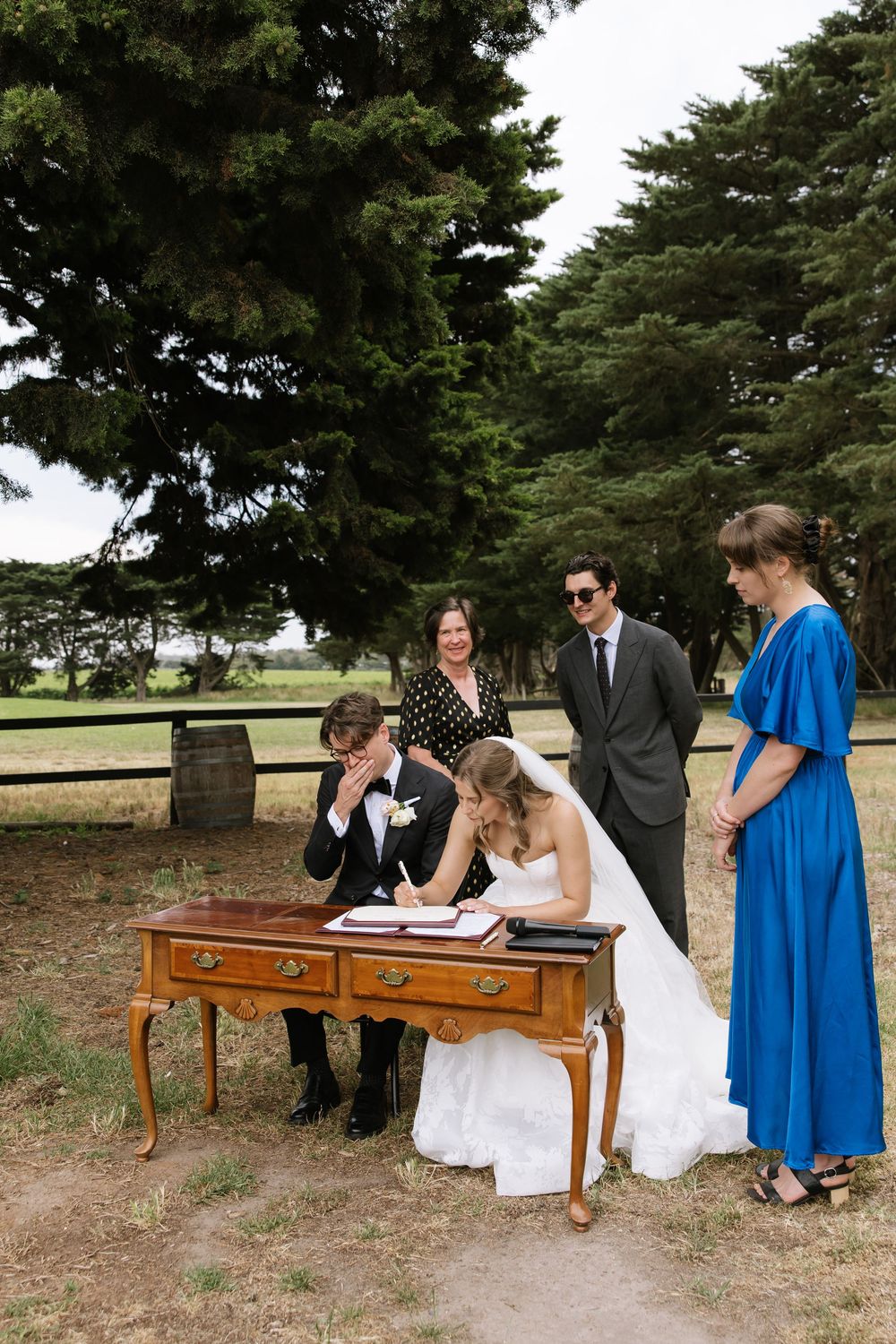 Bride and groom signing wedding register at outdoor ceremony with witnesses beside a wooden desk surrounded by trees.