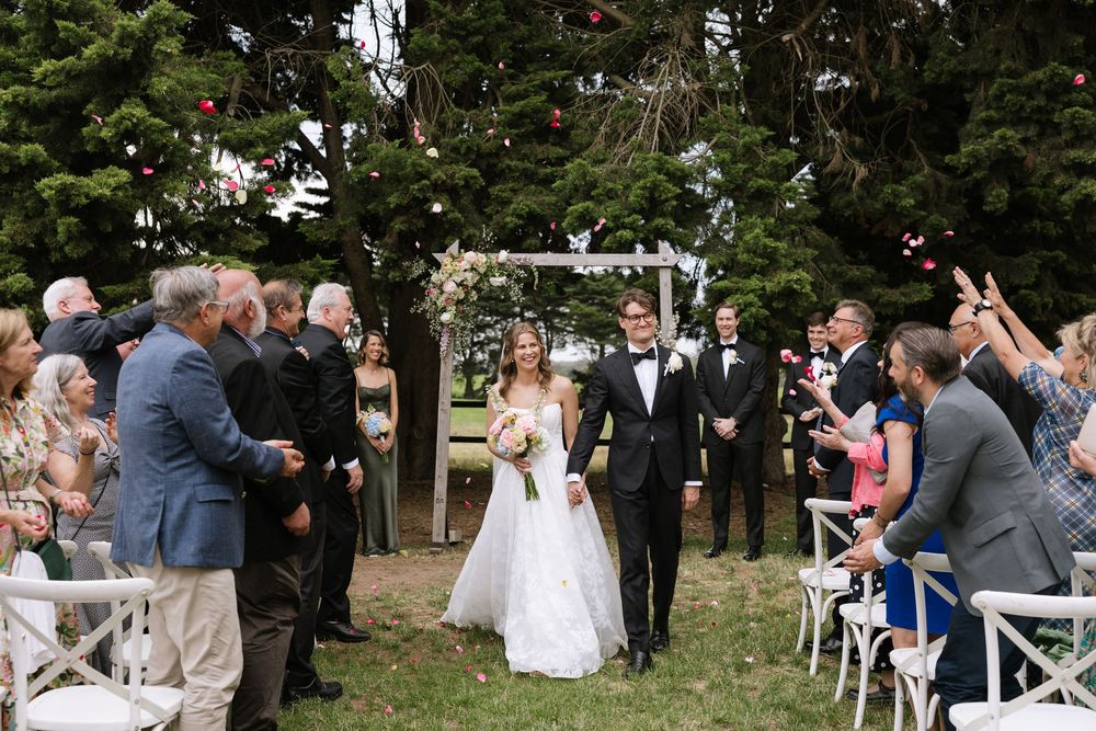 Bride and groom walk down outdoor aisle after wedding ceremony, guests cheering under floral arch surrounded by tall trees.