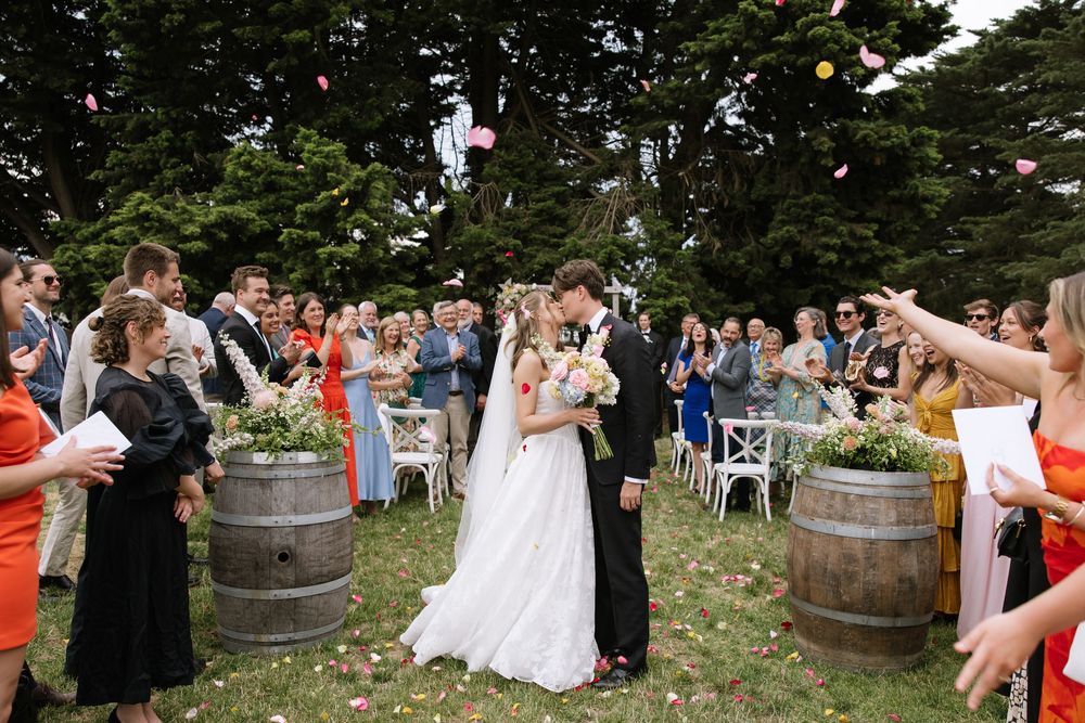 Bride and groom kiss while guests toss flower petals during outdoor wedding ceremony with barrel decorations.