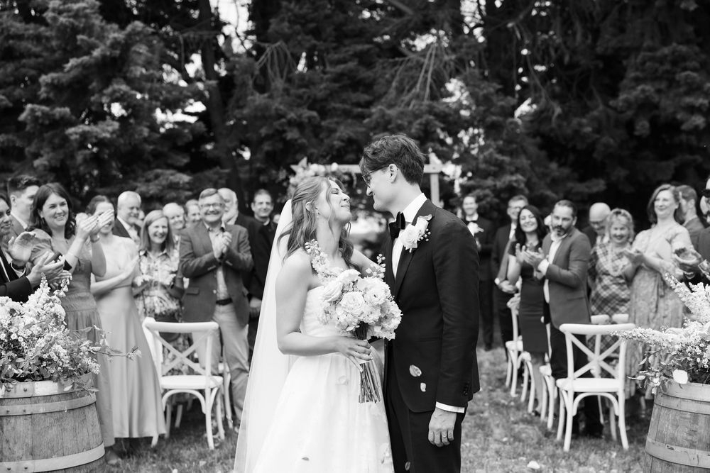 Black and white photo of a bride and groom sharing their first kiss at an outdoor wedding ceremony with guests cheering.
