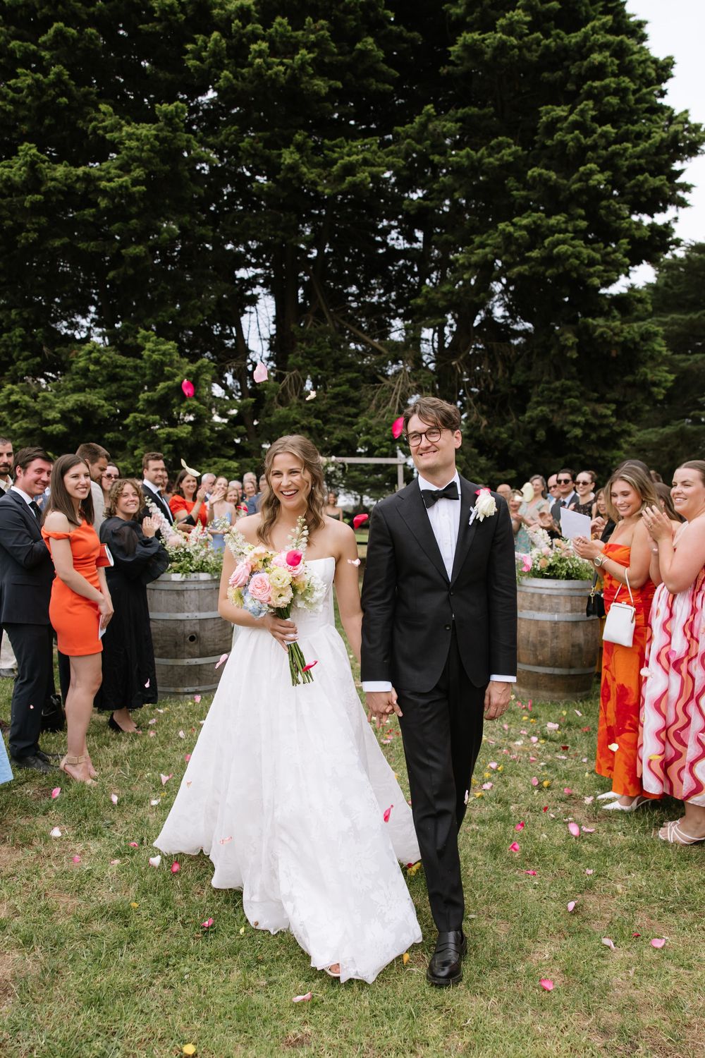 Bride and groom walk down the aisle as guests toss flower petals at an outdoor wedding ceremony.