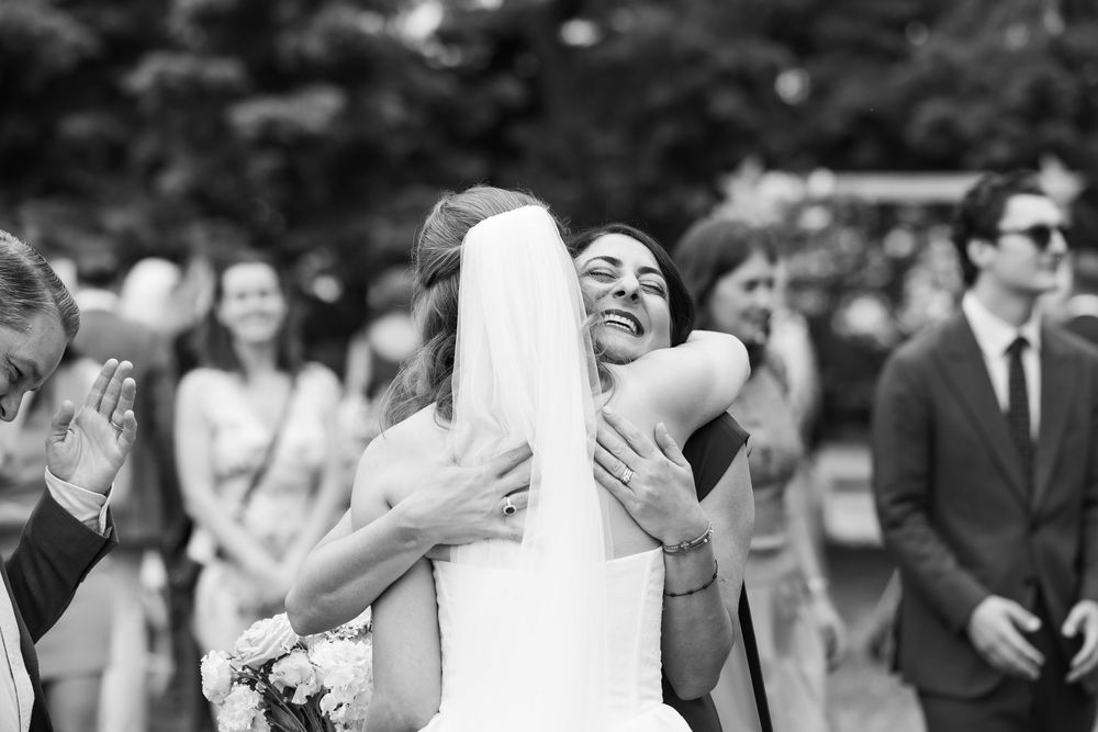 Bride in white gown shares joyful hug with guest at outdoor wedding ceremony, guests smiling in background.