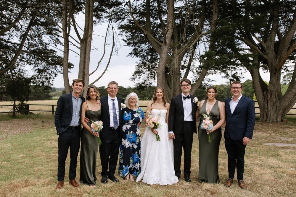 Wedding party of seven posing outdoors near large trees on a sunny day, featuring bride in white gown and groom in tuxedo.